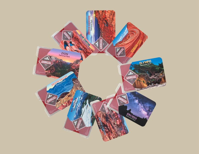May include: A collection of annual National Park passes, each featuring a different park scene. The passes are in clear plastic holders and display park names like Zion, Arches, and Olympic. The word "ANNUAL" is printed on each pass.