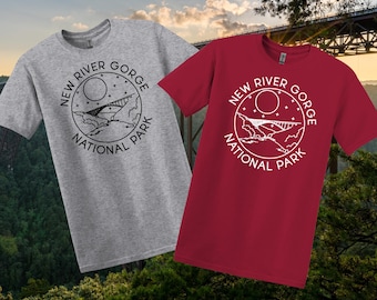 May include: Two t-shirts, one grey and one red, each displaying a circular graphic with the text "NEW RIVER GORGE NATIONAL PARK." The graphic includes a bridge, moon, stars, and landscape elements. The shirts are short-sleeved.