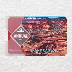 May include: A Grand Canyon National Park annual pass in a clear plastic holder. The pass features a colorful illustration of the canyon and the words "Annual Pass" in a diamond-shaped logo. The words "Grand Canyon National Park" are also visible.