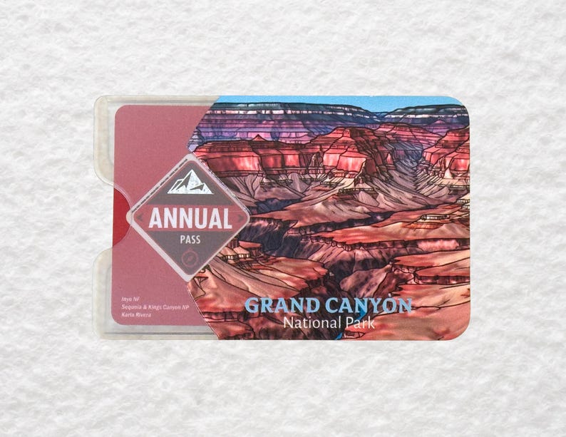 May include: A Grand Canyon National Park annual pass in a clear plastic holder. The pass features a colorful illustration of the canyon and the words "Annual Pass" in a diamond-shaped logo. The words "Grand Canyon National Park" are also visible.