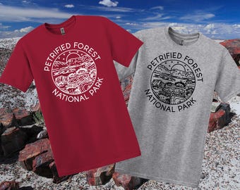May include: Two t-shirts, one red and one grey, each with a crew neck and the words "Petrified Forest National Park" in a circular design. The design features a landscape with a sun, mountains, and trees.