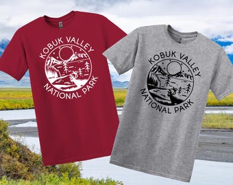 May include: Two short-sleeved t-shirts, one burgundy and one grey, each with a circular graphic. The graphic includes the text "KOBUK VALLEY NATIONAL PARK" around a mountain, lake, and deer scene. The shirts are displayed against a scenic background.