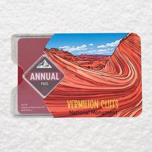 May include: A clear plastic holder containing a maroon Annual Pass card. The card features a landscape illustration of red rock formations under a blue sky, with the text "VERMILION CLIFFS National Monument."