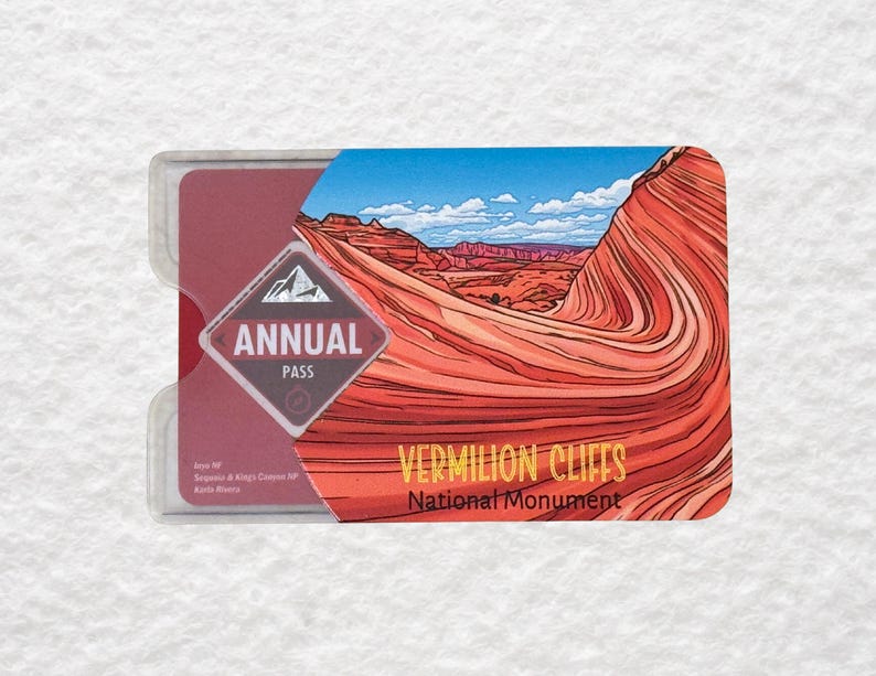 May include: A clear plastic holder containing a maroon Annual Pass card. The card features a landscape illustration of red rock formations under a blue sky, with the text "VERMILION CLIFFS National Monument."