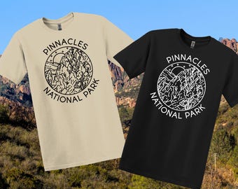May include: Two t-shirts, one beige and one black, each displaying a circular graphic with the text "PINNACLES NATIONAL PARK." The graphic shows a mountain range, sun, and trees. The shirts are short-sleeved and likely made of cotton.