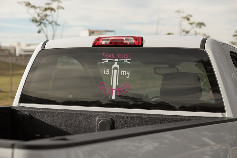 Puede incluir: Camioneta blanca con una calcoman&iacute;a rosa y blanca en la ventana trasera que dice "Trail Dust is my Glitter" con una silueta de una bicicleta.