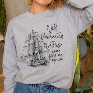 May include: A light gray sweatshirt featuring a detailed black and white illustration of a tall ship. The text "Wild Uncharted Waters come find me again" is printed above the ship. The sweatshirt has long sleeves and a crew neck.
