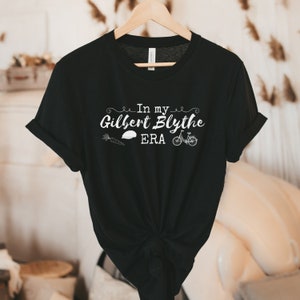 May include: Black t-shirt with white text that reads "In my Gilbert Blythe ERA" with a carrot, a hat, and a bicycle graphic.