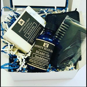 May include: A gift box filled with beard care products. The box contains a white bar of goat's milk soap, a bottle of sandalwood beard oil, and a black comb. The soap and oil are labeled "Infinity Blue Shop" and the soap is labeled "Black Sea".