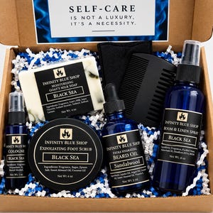 May include: A cardboard gift box containing Infinity Blue Shop products: a 4 oz goat's milk soap, a 1 oz cologne spray, a 4 oz exfoliating foot scrub, a 2 oz sandalwood beard oil, a room and linen spray, and a black comb. Text reads: SELF-CARE IS NOT A LUXURY, IT'S A NECESSITY.