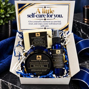 May include: A self-care gift box containing a bar of Infinity Blue Shop Black Sea goat's milk soap, a 4 oz container of exfoliating foot scrub, and a 1 oz cologne spray. The box lid features the text A little self-care for you.