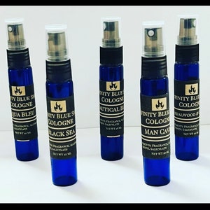 May include: Five small blue glass bottles with black labels and silver caps. Each bottle contains cologne and has a different scent name: Aqua Bleu, Black Sea, Nautical Blue, Man Cave, and Sandalwood & Oak. The bottles are arranged in a row on a white surface.