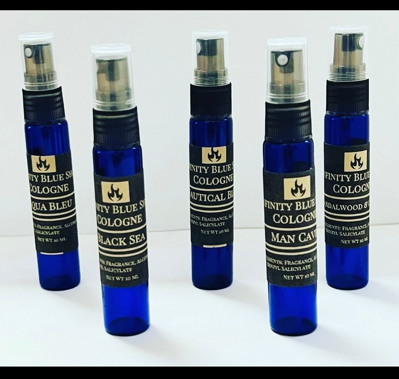 May include: Five small blue glass bottles with black labels and silver caps. Each bottle contains cologne and has a different scent name: Aqua Bleu, Black Sea, Nautical Blue, Man Cave, and Sandalwood & Oak. The bottles are arranged in a row on a white surface.