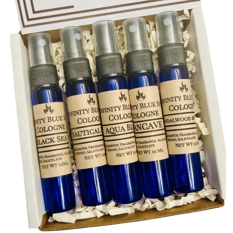 May include: Set of five small blue glass bottles with brown labels. The labels have white text that reads "Infinity Blue Cologne" followed by a scent name: "Black Sea", "Nautical", "Aqua Blanccave", "Sandalwood & Sea Salt", and "Black Sea". Each bottle has a spray nozzle and contains 10 ml of cologne.