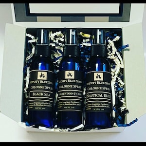 May include: A set of three blue glass cologne spray bottles with black labels. The labels have a gold flame logo and the text "Infinity Blue Shop Cologne Spray". The bottles are arranged in a white box with blue and white shredded paper. The scents are "Black Sea", "Sandalwood & Citrus", and "Nautical Bleu".