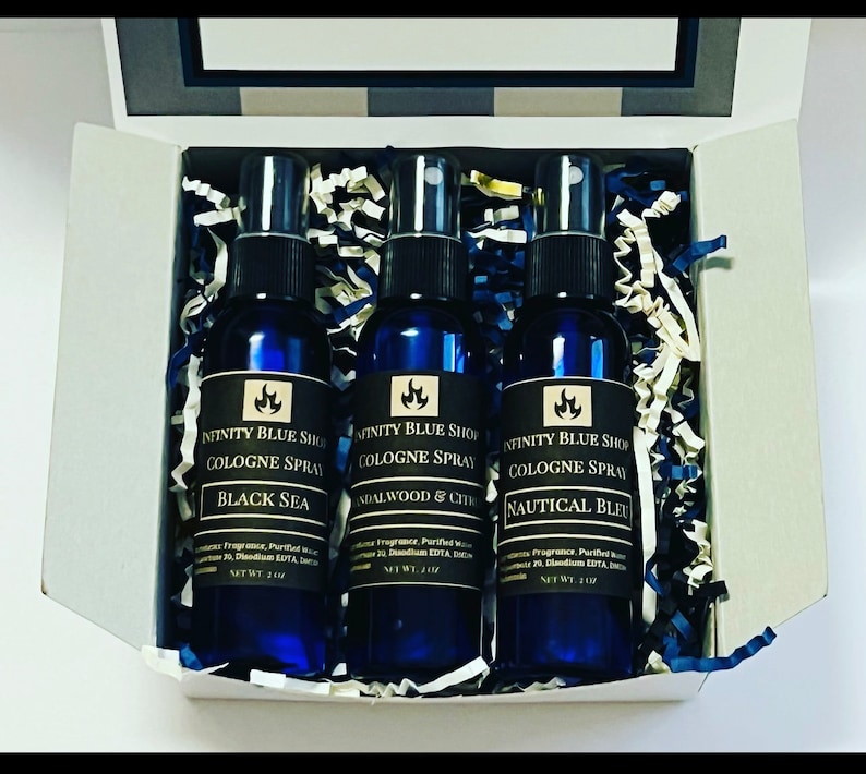 May include: A set of three blue glass cologne spray bottles with black labels. The labels have a gold flame logo and the text "Infinity Blue Shop Cologne Spray". The bottles are arranged in a white box with blue and white shredded paper. The scents are "Black Sea", "Sandalwood & Citrus", and "Nautical Bleu".
