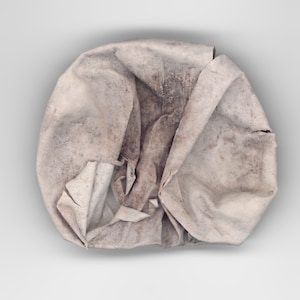 Könnte beinhalten: Ein kreisförmiges, cremeweißes Stoffstück mit einer stark zerknitterten und gefalteten Textur. Das Material weist dunkle, fleckige graue und braune Verfärbungen auf der Oberfläche auf, die ein abgenutztes Aussehen mit tiefen Schatten erzeugen.
