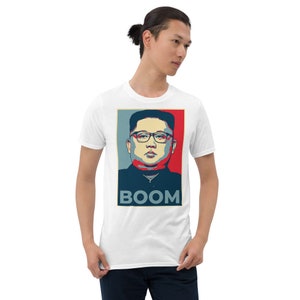 May include: White t-shirt with a graphic of a man wearing glasses and a red, white, and blue background. The text "BOOM" is printed below the graphic.
