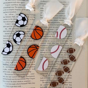May include: A close-up of a book with a white bookmark. The bookmark has a variety of sports ball designs, including a baseball, basketball, football, and soccer ball. The book has text on the pages, including the words "So... tell me... said" and "How did you get you".
