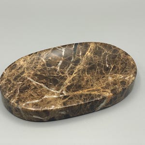 May include: Oval-shaped soap dish made of brown marble with white veining. The dish has a smooth, polished surface and a slightly raised edge. The marble pattern gives it a natural, elegant look.