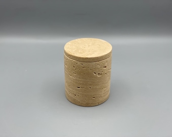 May include: A cylindrical, beige stone container with a matching lid. The container has a textured, layered appearance. The lid sits flush on top, creating a seamless look. The item is suitable for storage.