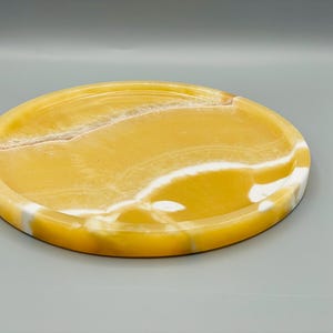 May include: A round, yellow and white marble tray with a smooth, polished surface. The tray has a natural, organic pattern of white veins running through the yellow marble.