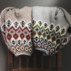 以下が含まれることがあります： 茶色、灰色、白、緑、赤の色合いの幾何学模様が施されたニットセーター2枚。セーターは折り畳まれ、木製の椅子にかけられています。