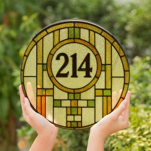 Op de afbeelding: Rond gebrandschilderd glazen huisnummerbord met het nummer 214 in een centrale cirkel. Het bord heeft een geel, groen en bruin kleurenschema met een zwarte rand. Het bord wordt door een persoon omhoog gehouden.