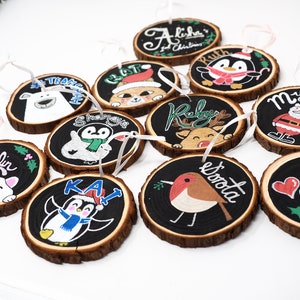 Peut inclure: Douze décorations en bois peintes à la main sur le thème de Noël. Les décorations représentent des pingouins, des rennes, le Père Noël, un rouge-gorge, un chat et un ours polaire. Chaque décoration porte un nom, comme "Ruby", "Kai" et "Mylan".