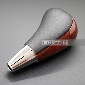 May include: A brown wood and black leather gear shift knob with a chrome tip. The knob has a textured leather surface and a wood grain pattern.