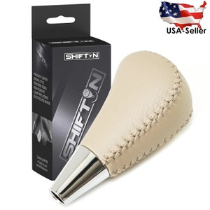 May include: Beige leather gear shift knob with a chrome base. The knob has a stitched design and is packaged in a box with the brand name "Shift'n".