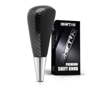 May include: A gear shift knob featuring a carbon fiber pattern on the sides, a black textured grip on the front, and a polished silver metallic base. Next to it stands a black rectangular box labeled SHIFTN PREMIUM SHIFT KNOB.