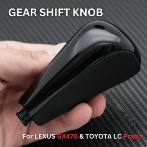 May include: A black gear shift knob with a glossy top and a textured matte side. The text GEAR SHIFT KNOB is at the top, and the text For LEXUS GX470 & TOYOTA LC Prado is at the bottom. The knob has a small cylindrical base attachment point.