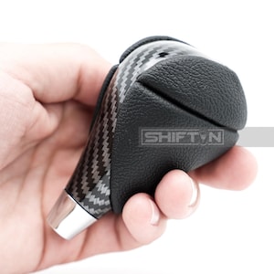 May include: A black and silver car gear shift knob with a carbon fiber pattern and a silver base. The knob has a textured leather grip and the text "SHIFT N" is visible.
