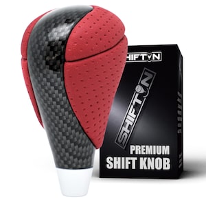 May include: A gear shift knob featuring a red perforated texture on the sides and a black carbon fiber pattern down the center. The knob is positioned in front of a black box with the text SHIFTON PREMIUM SHIFT KNOB printed in white.