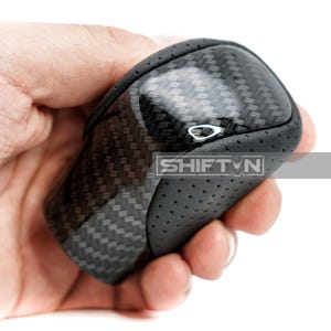 May include: A black and gray carbon fiber shift knob with the text "SHIFT N" on the side. The shift knob has a glossy top and a perforated leather-like grip.