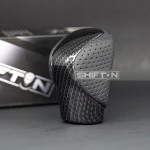 May include: A black and gray shift knob with a carbon fiber pattern and perforated leather top. The text "SHIFT N" is visible on the side. The knob is designed for a vehicle's gear shift.