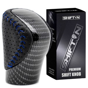 May include: A black and carbon fiber shift knob with blue stitching. The shift knob has a leather grip and is next to a black box with the text "SHIFT N PREMIUM SHIFT KNOB".