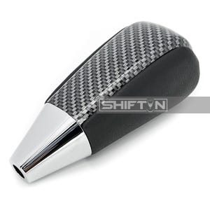 May include: A car gear shift knob with a carbon fiber weave top and black leather sides. The bottom is chrome with the text "SHIFT N" in a white rectangle.