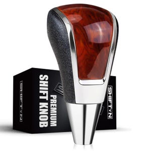 May include: A gear shift knob featuring a polished wood grain top insert, a textured black side grip, and a smooth silver metallic base. The knob is positioned in front of a black rectangular box with the white text PREMIUM SHIFT KNOB and SHIFT N.
