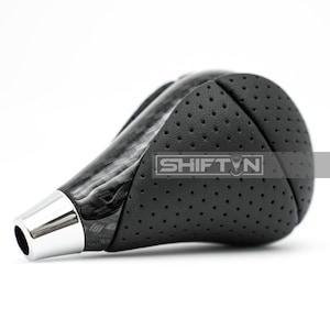 May include: A black and silver shift knob with a carbon fiber pattern and perforated leather grip. The shift knob has a chrome base and the text "SHIFT N" is visible on the side.