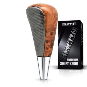 May include: A gear shift knob featuring a polished chrome base, a textured dark gray carbon fiber-style front panel, and a smooth brown burl wood-grain back. Next to it is a black box labeled SHIFTON PREMIUM SHIFT KNOB with a gear pattern logo.