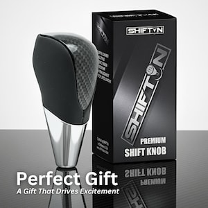 May include: A black and silver shift knob with a carbon fiber top, next to a black box with "SHIFT N" and "PREMIUM SHIFT KNOB" printed on it. The text "Perfect Gift" and "A Gift That Drives Excitement" are also visible.