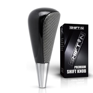 May include: A black gear shift knob with a textured gray front panel and a polished silver base, displayed next to a black product box labeled SHIFTON PREMIUM SHIFT KNOB with white text and a graphic of a gear pattern on the letter O.