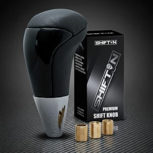 May include: A black and silver shift knob with a leather top and a glossy black front. The product is next to a black box with the text "SHIFTYN" and "PREMIUM SHIFT KNOB". Three gold-colored bushings are also included.