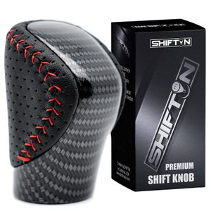 May include: A gear shift knob featuring a black carbon fibre pattern and perforated black side panels with red stitching. Next to it is a black rectangular box labelled SHIFTIN PREMIUM SHIFT KNOB with a graphic of a gear shifter and text.