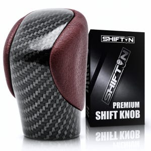 May include: A gear shift knob featuring a central carbon fiber pattern panel flanked by textured maroon leather sides. Next to it stands a black rectangular box with the text SHIFTON and PREMIUM SHIFT KNOB printed in white lettering.