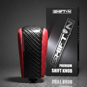 May include: A carbon fiber and red leather shift knob with the text "SHIFT N" on the packaging. The shift knob has a black and gray checkered pattern and red leather sides. The packaging reads "PREMIUM SHIFT KNOB".