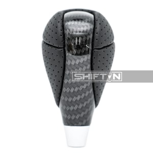 May include: A black and gray shift knob with a carbon fiber pattern and the text "SHIFT N". The top portion is perforated black leather. The base is white.