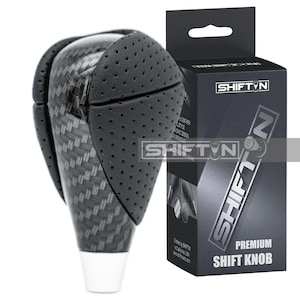 May include: A black and carbon fiber shift knob with a perforated leather grip. The shift knob is next to a black box with the text "SHIFTN" and "PREMIUM SHIFT KNOB".
